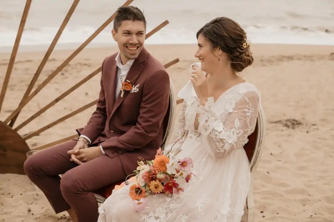 La mariée en robe de dentelle et le marié en costume bordeaux, sur la plage